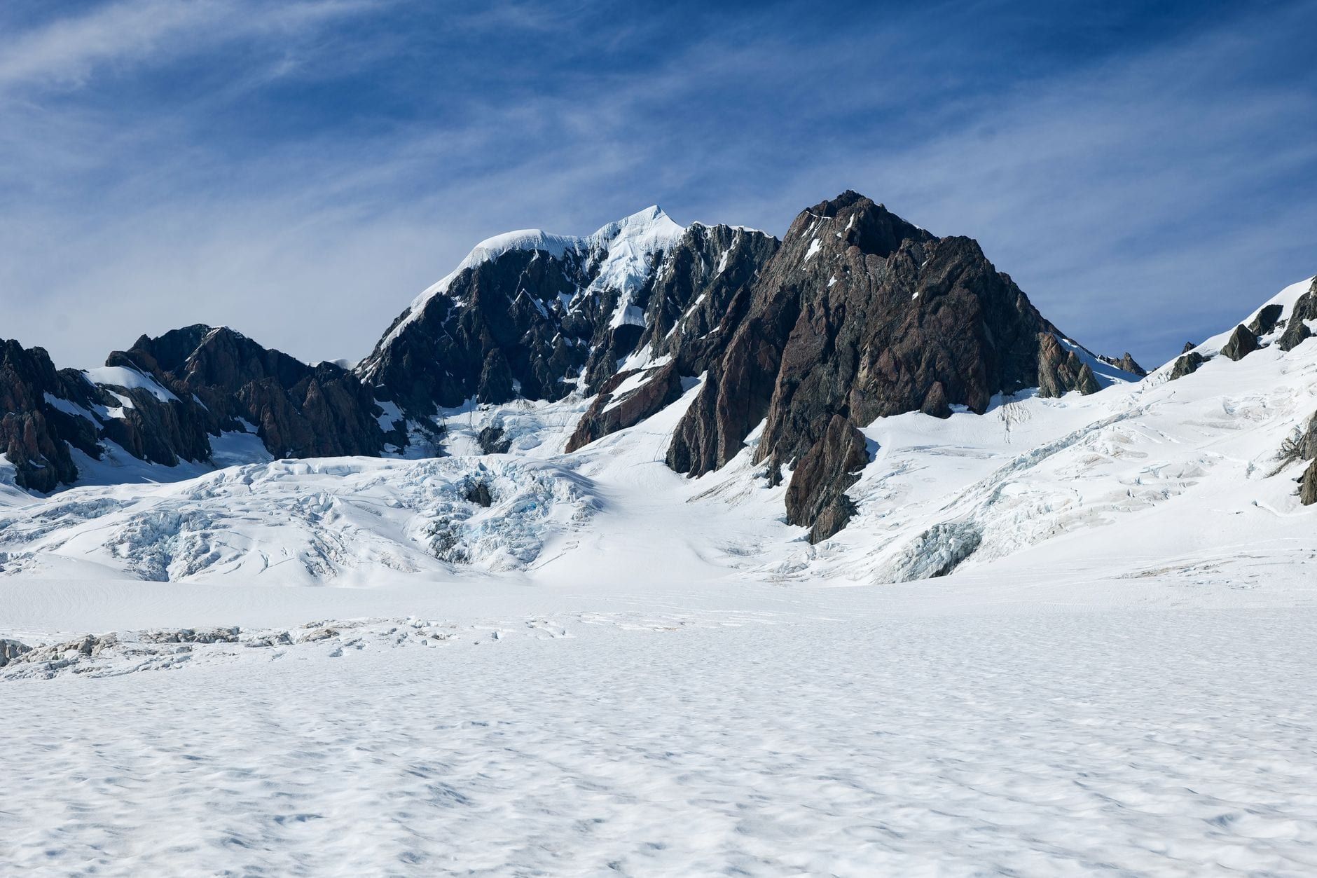 Snow and ice stretch across Fox Glacier beneath rugged mountain peaks under a clear blue sky.