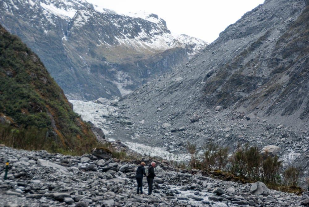 Fox Glacier's pale ice terminal face lies at the head of a rugged mountain valley, seen across a rocky riverbed with hikers in the foreground.