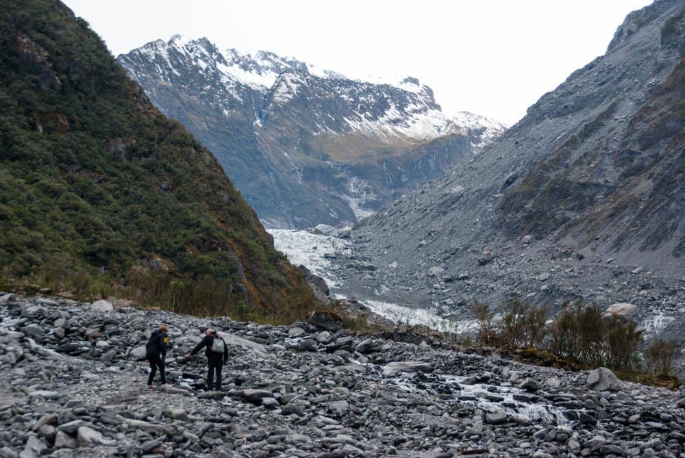 Hikers walk across a rocky riverbed toward Fox Glacier framed by steep mountain slopes and snowy peaks.