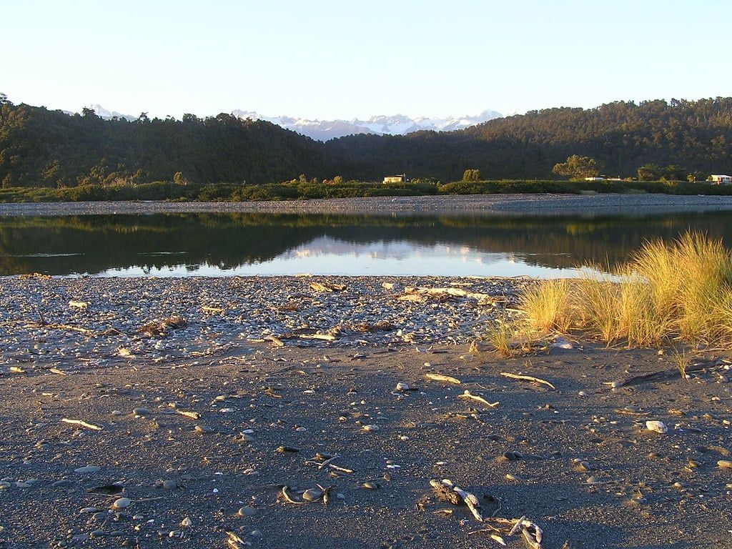 Calm waters of Okarito Lagoon reflect forested hills with snowcapped peaks of the Southern Alps in the distance near Fox Glacier.
