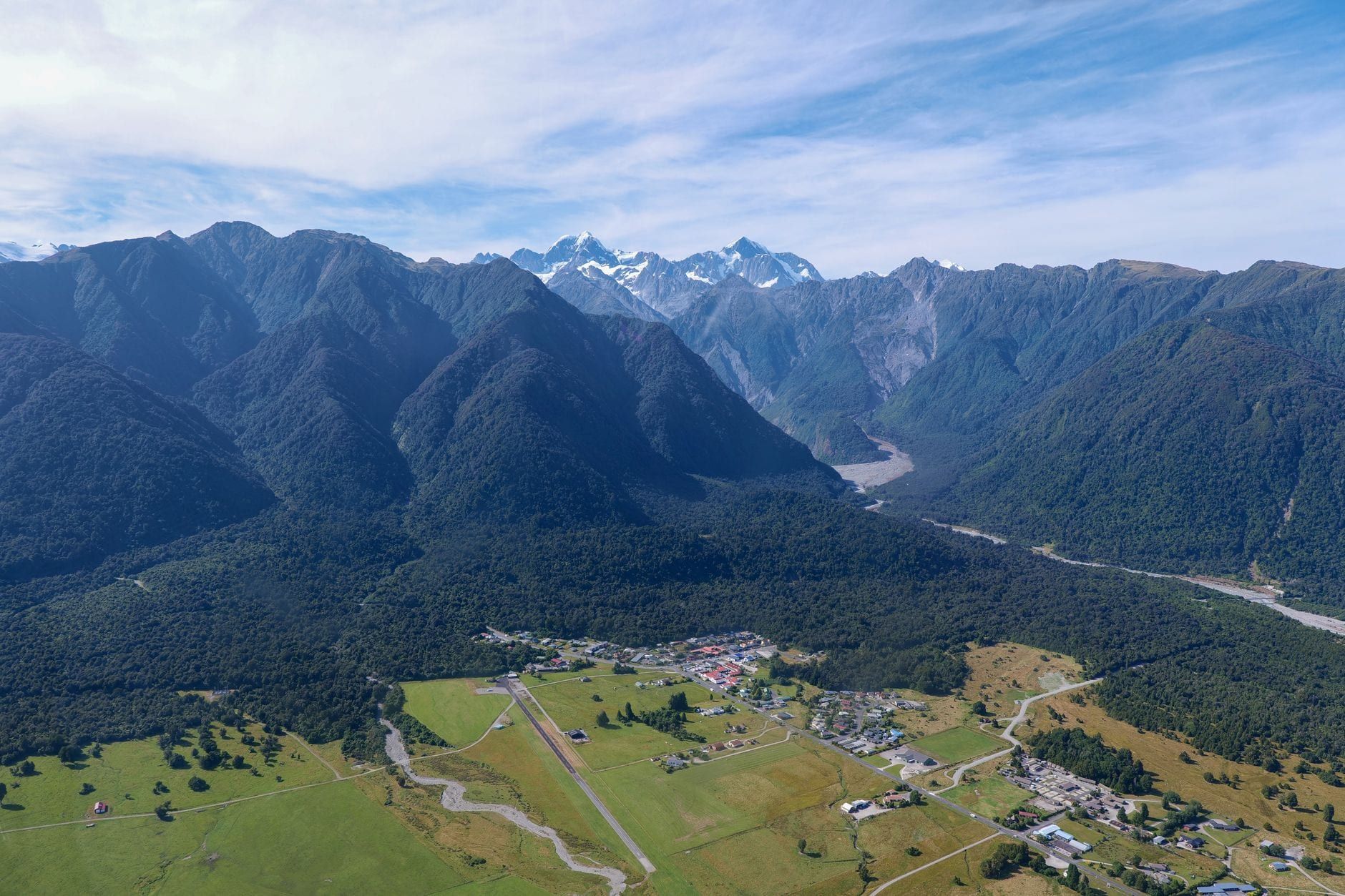 Aerial view of Fox Glacier Village set against lush green pastures, dense native forest, and the snow-capped Southern Alps.
