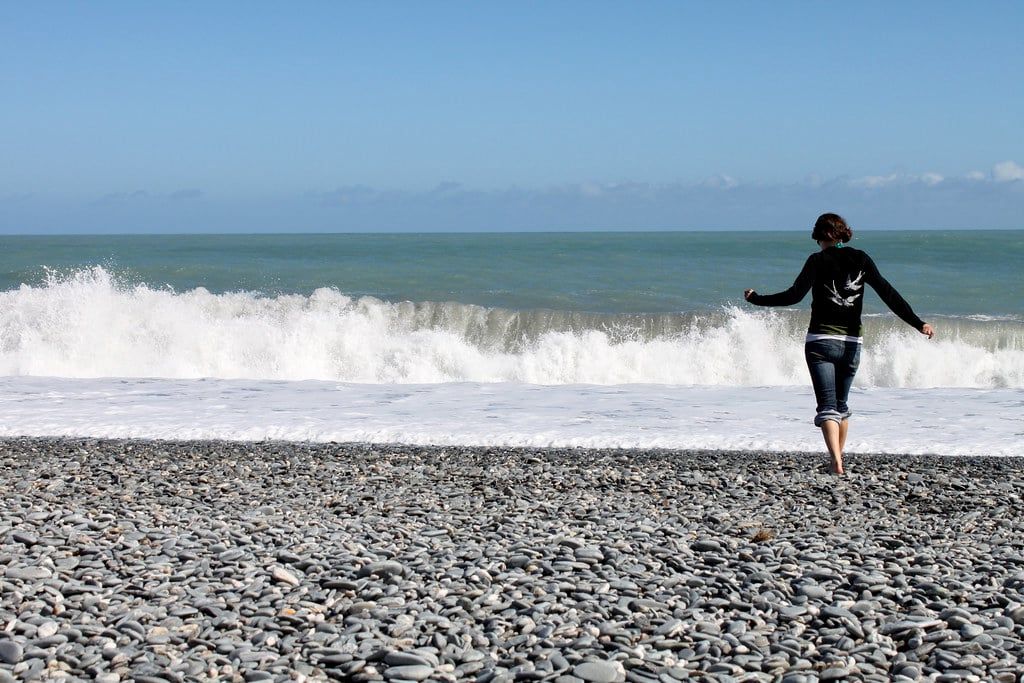 A person walks across the pebble shoreline toward breaking waves at Gillespies Beach near Fox Glacier.
