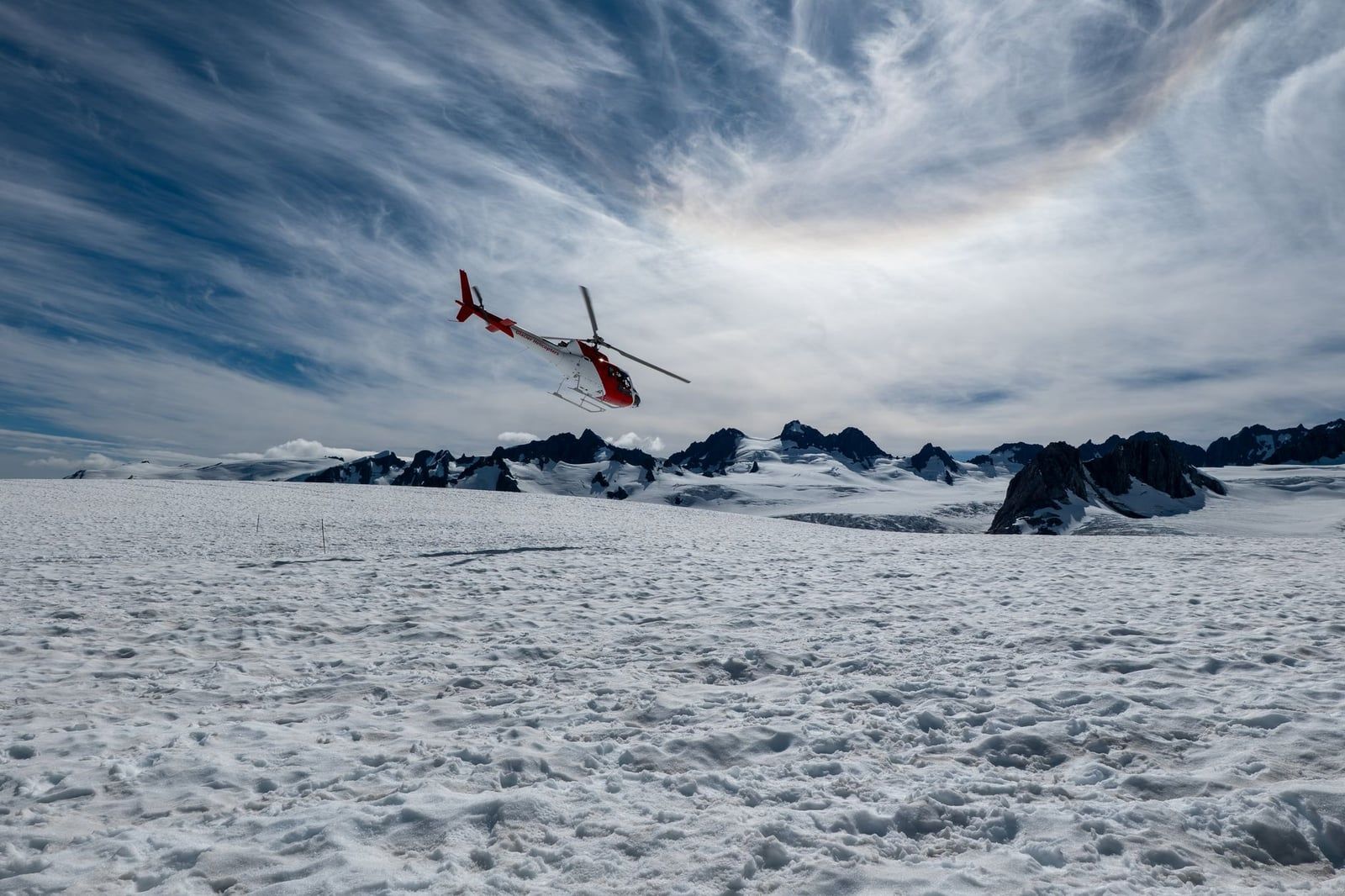 A red and white helicopter flies low over the snow-covered expanse of Fox Glacier with jagged mountain peaks in the background.